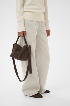 Persona valkā krēmkrāsas platu kāju bikses un džemperi, turot tumši brūnu zamšādas rokassomiņu, kas saskaņota ar tumši brūnām dzīvokļveida kurpēm.