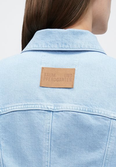 Ανοιχτό μπλε τζιν μπουφάν με μυτερό γιακά, ραφές σε αντίθεση και ορθογώνια καφέ δερμάτινη ετικέτα με ανάγλυφο κείμενο στην πλάτη.