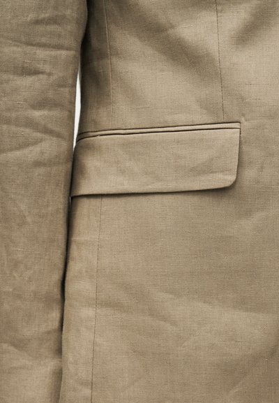 Κοντινό πλάνο μανσετών μπεζ λινής ζακέτας και τσέπης με καπάκι στο πλάι, που δείχνει την υφή του υφάσματος και τις λεπτομέρειες των ραφών.
