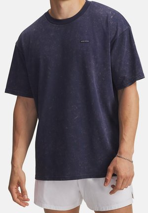 Homme portant un t-shirt Under Armour bleu marine foncé à manches courtes et un short blanc, avec un bracelet chaîne en argent à son poignet droit.