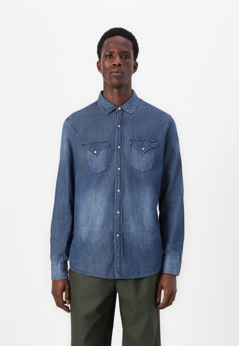 Camisa de denim en azul medio con botones en la parte delantera, dos bolsillos en el pecho y un cuello puntiagudo. Presenta una textura desgastada y botones en tono plateado.