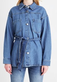 Chaqueta de mezclilla en azul claro con cierres de botón, dos bolsillos en el pecho y una cintura con cinturón. Cuenta con un cuello con solapas y mangas largas.