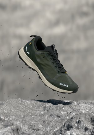 Zapatilla de trail running verde oscuro y beige con cordones negros, en el aire sobre un terreno rocoso, levantando pequeñas cantidades de tierra y escombros.