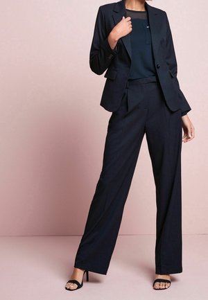 Femme portant un blazer noir cintré, un pantalon large assorti et des sandales à talons hauts noires ouvertes, debout devant un fond rose pâle.