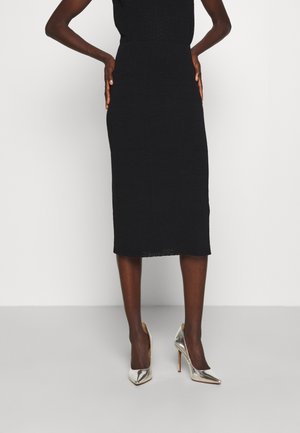 Personne portant une jupe midi noire cintrée et des chaussures à talons hauts pointues argentées métalliques, debout avec les mains sur les hanches.