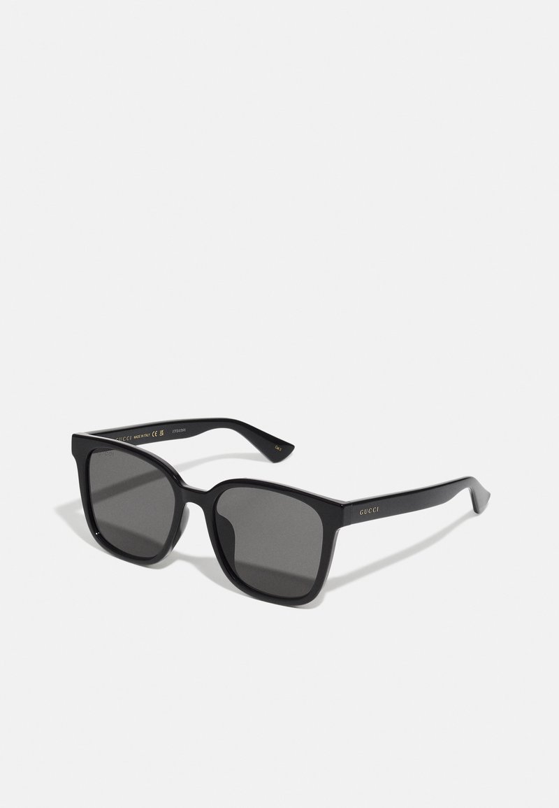 Gafas de sol negras con lentes cuadrados y un marco de plástico brillante. Incluyen un logo dorado en las patillas y lentes de degradado oscuro.