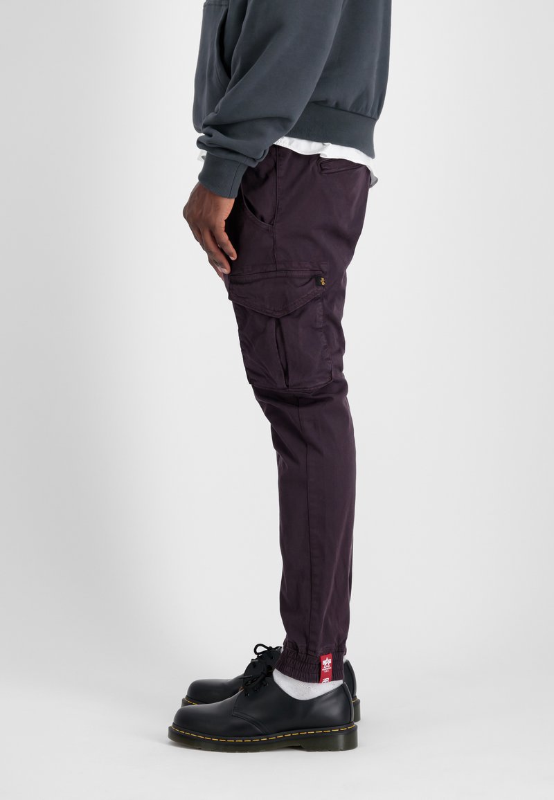 Fioletowe spodnie cargo z wieloma kieszeniami i elastycznymi mankietami, w zestawieniu z czarnymi butami o grubych podeszwach i żółtymi przeszyciami.