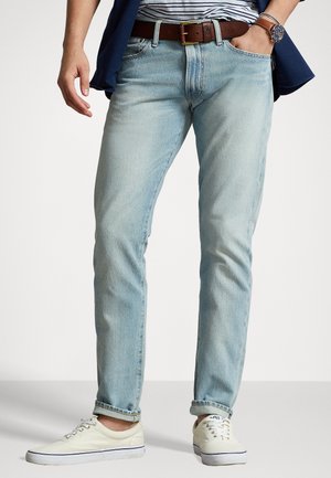 Mann trägt hellblaue Jeans mit aufgerollten Säumen, weiße Canvas-Sneaker, braunen Gürtel, gestreiftes Hemd, marineblaue Jacke und Armbanduhr.