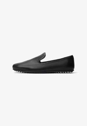 Zapato negro de cuero sin cordones con una textura suave, puntera ovalada y costuras mínimas. Presenta una suela de goma con pequeños protuberancias para mayor agarre.