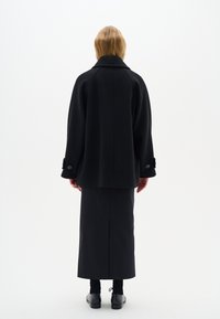 Zwarte wollen jas met een brede kraag, oversized pasvorm en knoopmanchetten, gecombineerd met een zwarte jurk die tot de vloer reikt en veterschoenen.
