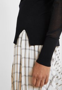Maglione nero a maglia con orlo a coste abbinato a una gonna plissettata beige con un motivo a quadretti in linee marroni scure. La texture è liscia e leggera.