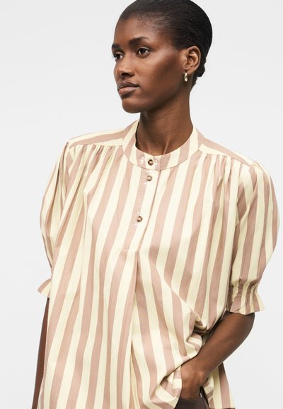 Γυναίκα με κοντά μαλλιά φοράει μπεζ και εκρού μπλούζα με κάθετες ρίγες και φουσκωτά μανίκια, καθώς και χρυσούς κρίκους στα αυτιά, κοιτάζοντας πλαγίως.
