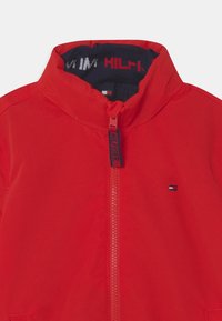 Rød jakke med høj krave og lynlås foran. Har en kontrast navy-farvet foring og et lille logo-patch på venstre side. Glat stoftekstur.