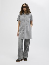 Vestido camisola de ganga cinza com mangas curtas, fecho de botão frontal e um bolso no peito, combinado com calças de ganga cinza de perna larga e botas pretas resistentes.