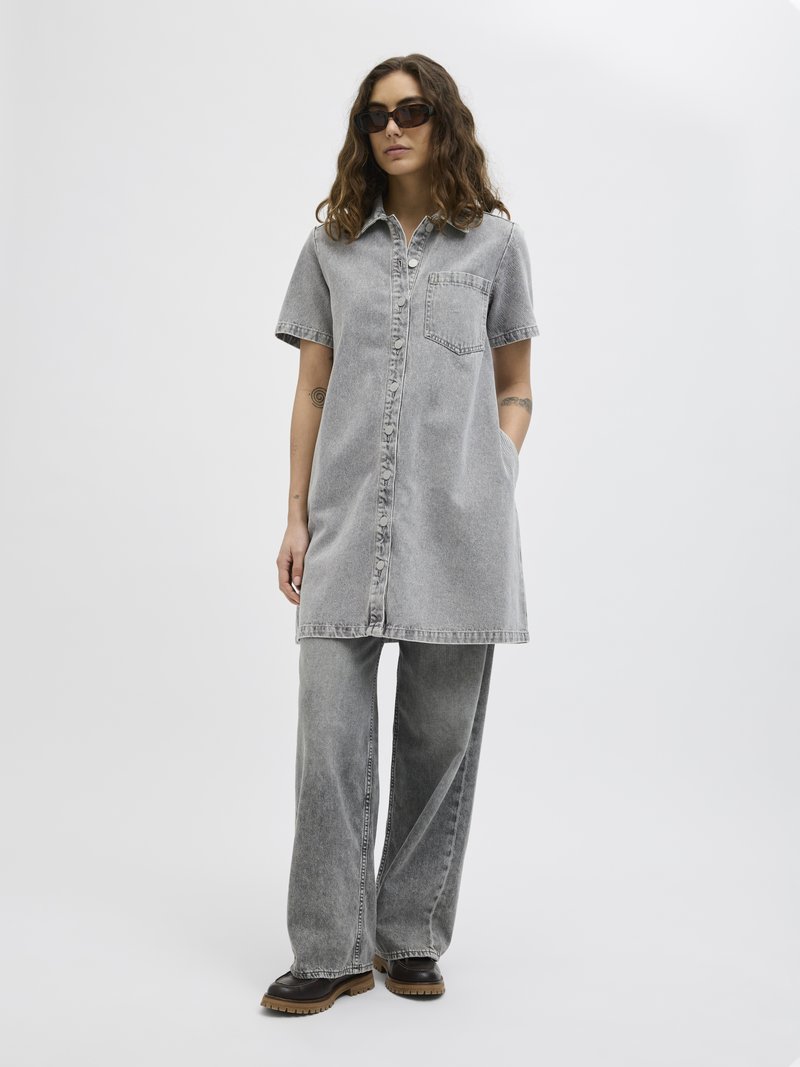 Vestido camisola de ganga cinza com mangas curtas, fecho de botão frontal e um bolso no peito, combinado com calças de ganga cinza de perna larga e botas pretas resistentes.