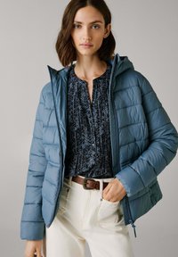 Vrouw in een blauwe gewatteerde hooded jas over een donker gebloemde blouse, met crèmekleurige pantalons en een bruine riem, poserend tegen een effen achtergrond.