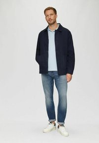Mann steht in blauen Jacke, hellblauem Polohemd, hochgerollten blauen Jeans und weißen Sneakers vor einem weißen Hintergrund.