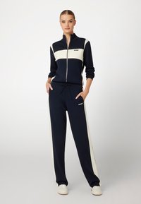 Jūrų kariuomenės zip-up treniruočių striukė su grietinėlės spalvos juostelėmis, derinama su atitinkančiomis kelnėmis, turinčiomis raištelių juosmenį ir logotipo detalę. Minkšta medžiaga, laisvas siluetas.
