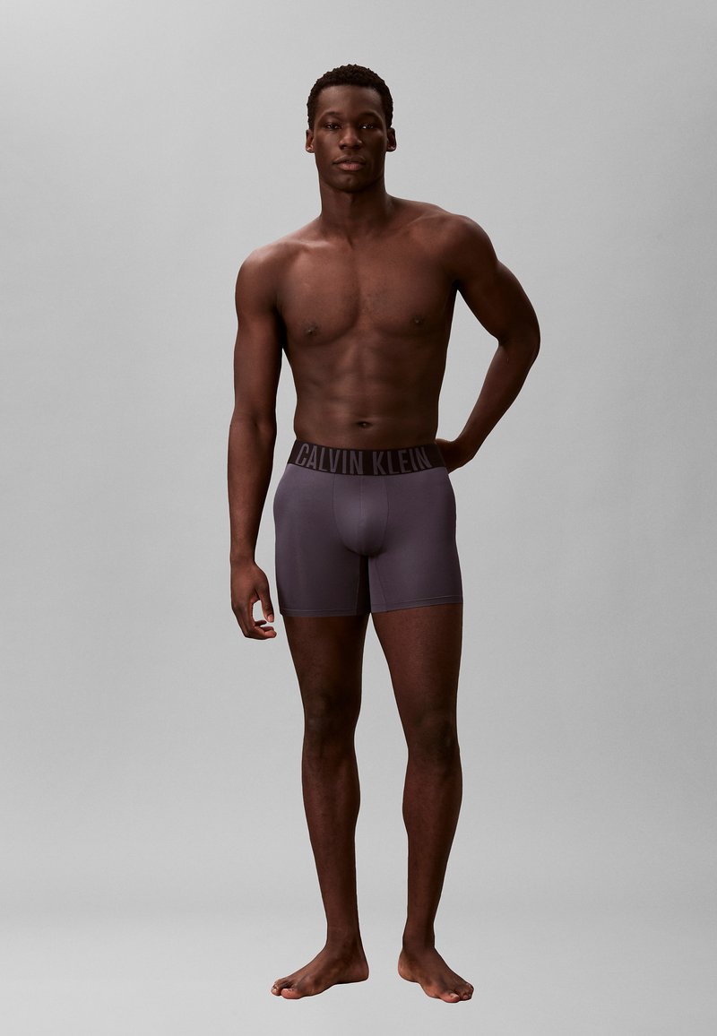 Jeune homme noir debout pieds nus, portant un caleçon ajusté Calvin Klein gris foncé, une main reposant sur sa hanche.