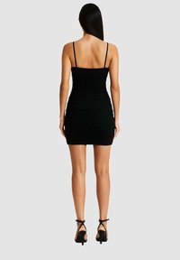 Donna con capelli lunghi e lisci che indossa un vestito mini nero arricciato senza maniche e sandali neri con tacchi alti, in piedi di spalle.