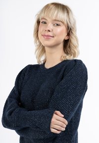 Sötétkék textúrált kötött pulóver hosszú ujjakkal, puffos vállrésszel és bordás mandzsettákkal, amit egy szőke hajú személy visel.
