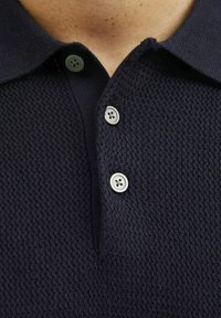 Tamsiai mėlynas tekstūruotas polo marškinėliai su apykakle ir trimis šviesių spalvų mygtukais prie įėjimo. Artimas vaizdas.