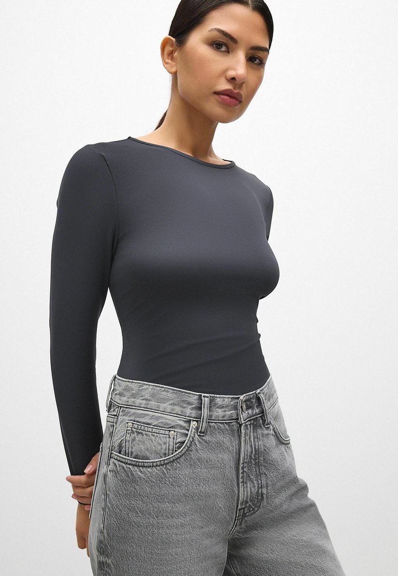 Langsleeve top in donkergrijs, gemaakt van gladde stof. Heeft een ronde hals en een aansluitend ontwerp, gecombineerd met lichtgrijze hooggetailleerde jeans.