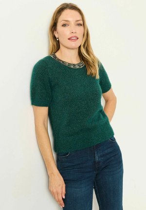Kvinde stående med den ene hånd i lommen iført en grøn kortærmet sweater med perlepyntet halsudskæring og blå jeans mod hvid baggrund.