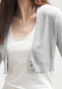 Světlově šedý zkrácený kardigan z jemného pleteného materiálu s vroubkovanými manžetami a knoflíkovým detailem vpředu, nošený přes bílou košili.