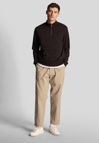 Junger Mann steht mit den Händen in den Taschen, trägt einen schwarzen Viertelreißverschluss-Pullover, beigefarbene Hose, weiße Sneaker und weiße Socken vor grauem Hintergrund.