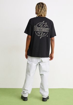 Camiseta de algodón negra con un gran gráfico en blanco y amarillo de "Detroit" en la parte posterior, combinada con pantalones cargo holgados en gris claro.