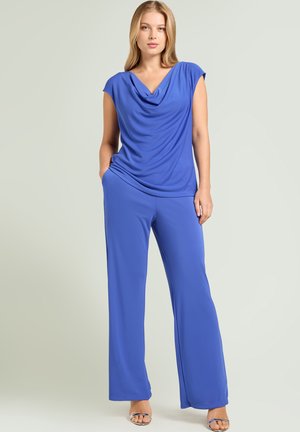 Kobieta stojąca z częściowo wsuniętymi dłońmi do kieszeni, ubrana w niebieski, bezrękawnik z drapowanym przodem oraz pasujące szerokie spodnie, do tego srebrne sandały.