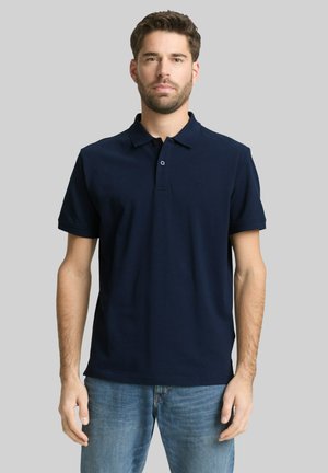Homme portant un polo bleu marine à manches courtes et un jean bleu, debout devant un fond gris clair uni.
