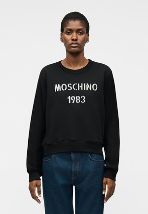 Γυναίκα που φοράει μαύρο φούτερ με λευκή επιγραφή «MOSCHINO 1983» και μπλε τζιν παντελόνι, στέκεται μπροστά σε απλό ανοιχτόχρωμο φόντο.