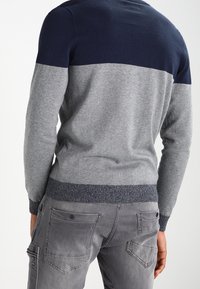 Světlě šedý svetr s tmavě modrou horní částí, s texturovanými pletenými manžetami a přiléhavým střihem, doplněný šedými kapsáčovými kalhotami s kapsami.