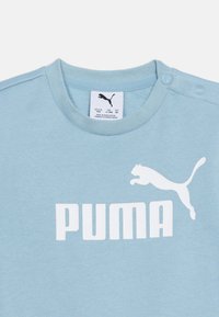 Ανοιχτό μπλε παιδικό πουκάμισο με λευκό λογότυπο Puma και όνομα της μάρκας, με δύο κουμπιά σταθερά στον ώμο για εύκολη φορεσιά.