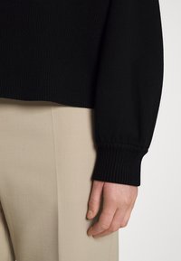 Pull en tricot noir avec des poignet côtelés, associé à un pantalon beige taillé. Gros plan sur une main reposant contre le pantalon, montrant la texture.