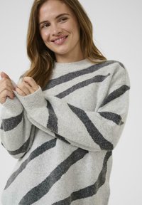 Femme avec de longs cheveux bruns souriant, portant un pull gris clair avec des rayures diagonales gris foncé, tenant le poignet du pull avec les deux mains.