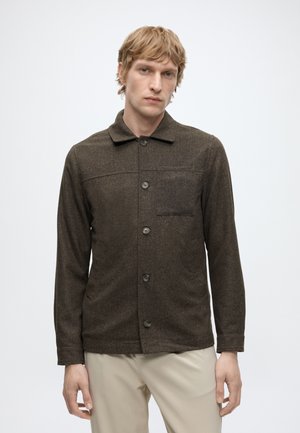 Ung mand med lyst hår iført en brun knappet jakke og beige bukser, der står mod en ensfarvet hvid baggrund.