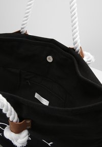 Borsa in tela nera con manici in corda bianca, dotata di chiusura a scatto metallica e una tasca interna con zip. L'etichetta riporta la scritta "Seafolly."