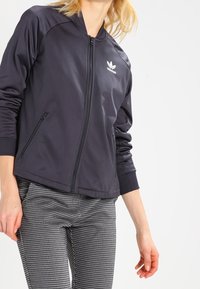 Svart zip-up jacka med en slät, lätt känsla, med en vit Adidas-logotyp på vänster bröst och två framfickor med dragkedja.