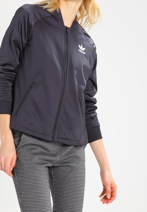 Czarna kurtka zapinana na zamek o gładkiej, lekkiej teksturze, z białym logo Adidas na lewym piersiu i dwoma przednimi kieszeniami na zamek.