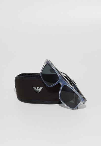 Ochelari de soare cu o ramă din plastic transparent și lentile întunecate, așezați pe un etui rigid negru cu un logo argintiu și închidere cu fermoar.