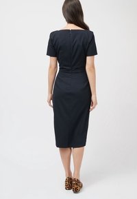 Navy-blaues, tailliertes Midikleid mit kurzen Ärmeln, quadratischem Ausschnitt, dezenten Reißverschluss im Rücken und glattem Stoff. Tragen mit gemusterten braunen Absätzen.
