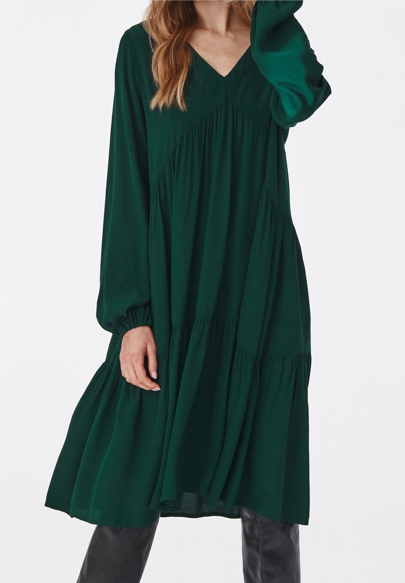 Robe verte à manches longues avec un col en V, présentant un design à étages et une jupe fluide, fabriquée en tissu doux avec une texture lisse.