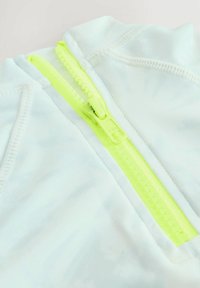 Tessuto azzurro chiaro con una texture liscia presenta una vivace cerniera verde neon all'altezza del collo, circondata da sottili dettagli di cucitura.