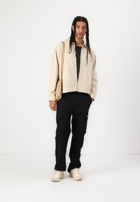 Beige striped jacket, black cargo pants, and light beige sneakers. Model holds a small tan shoulder bag, with braided hair visible.