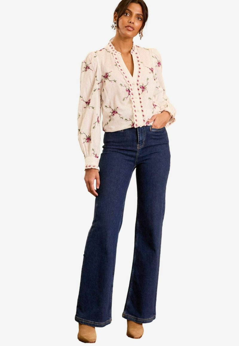 Blusa a fiori in crema con maniche a palloncino e scollatura a V, abbinata a jeans svasati blu scuro e calzature beige.