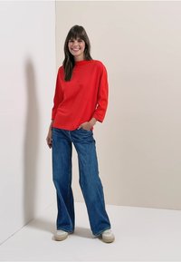 Rotes Langarmshirt mit Rundhalsausschnitt, kombiniert mit weit geschnittenen blauen Jeans und hellen Sneakers, vor einem neutralen Hintergrund stehend.
