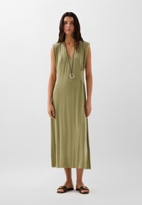 Abito lungo senza maniche di un verde oliva con scollatura a V profonda e arricciature laterali, abbinato a sandali marroni con un accento metallico decorativo.